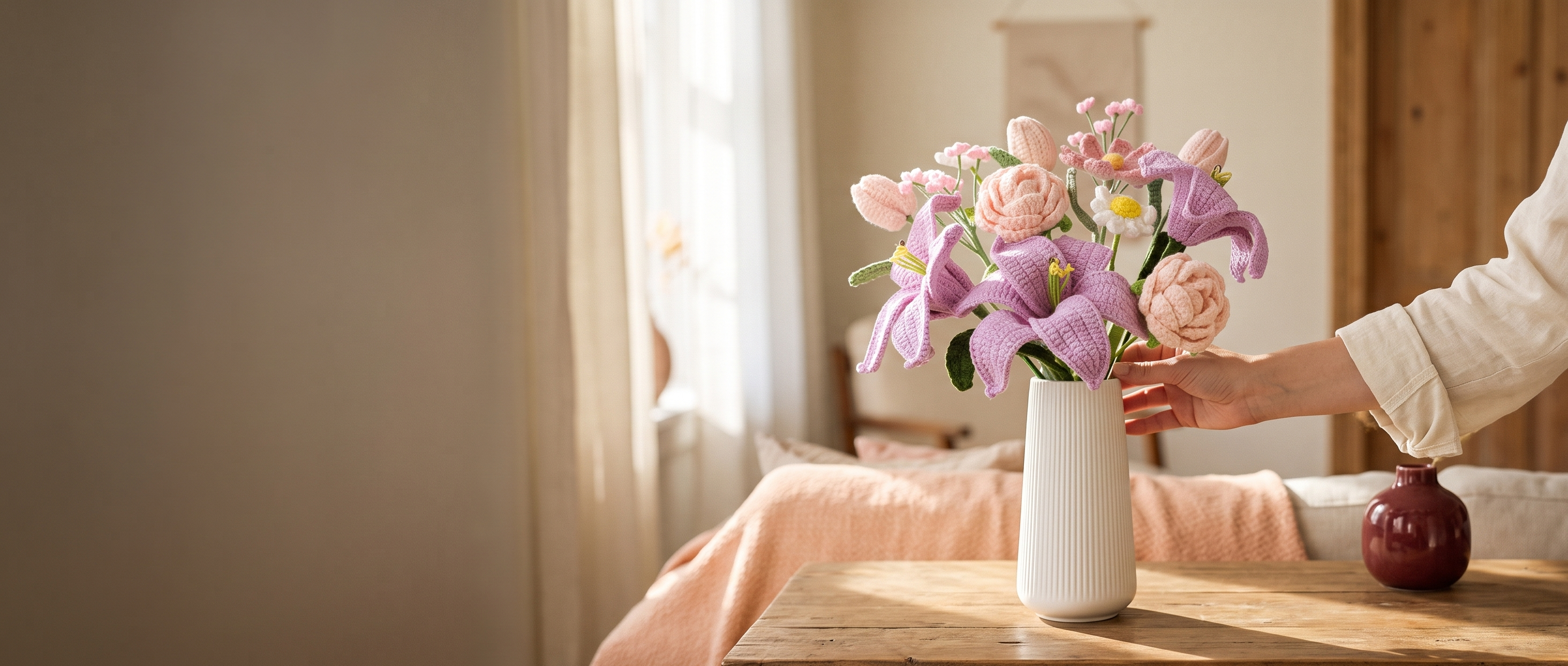 A person in a white long-sleeve top adjusts a bouquet of crochet flowers in a white ribbed vase on a wooden table in a softly lit bedroom.